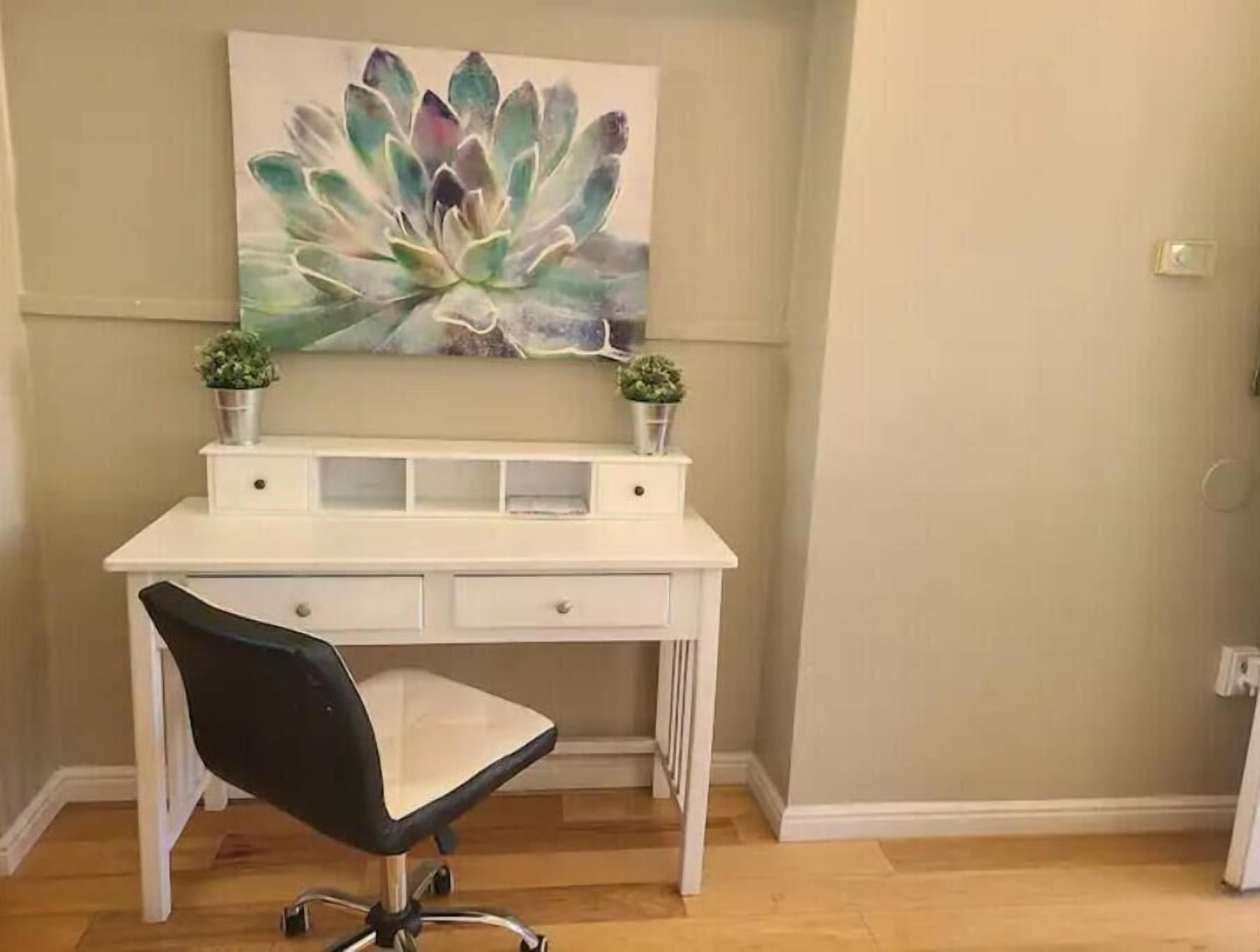 A white desk with two small potted plants, a black office chair, and a large abstract painting on the wall.