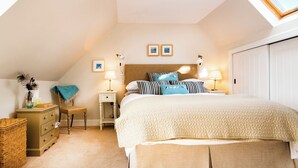 A bedroom with a sloped ceiling, a bed with a quilted cover, bedside lamps, a small table, a chair, a wicker basket, and a patterned rug. - White House - UKC6895 (Golspie)