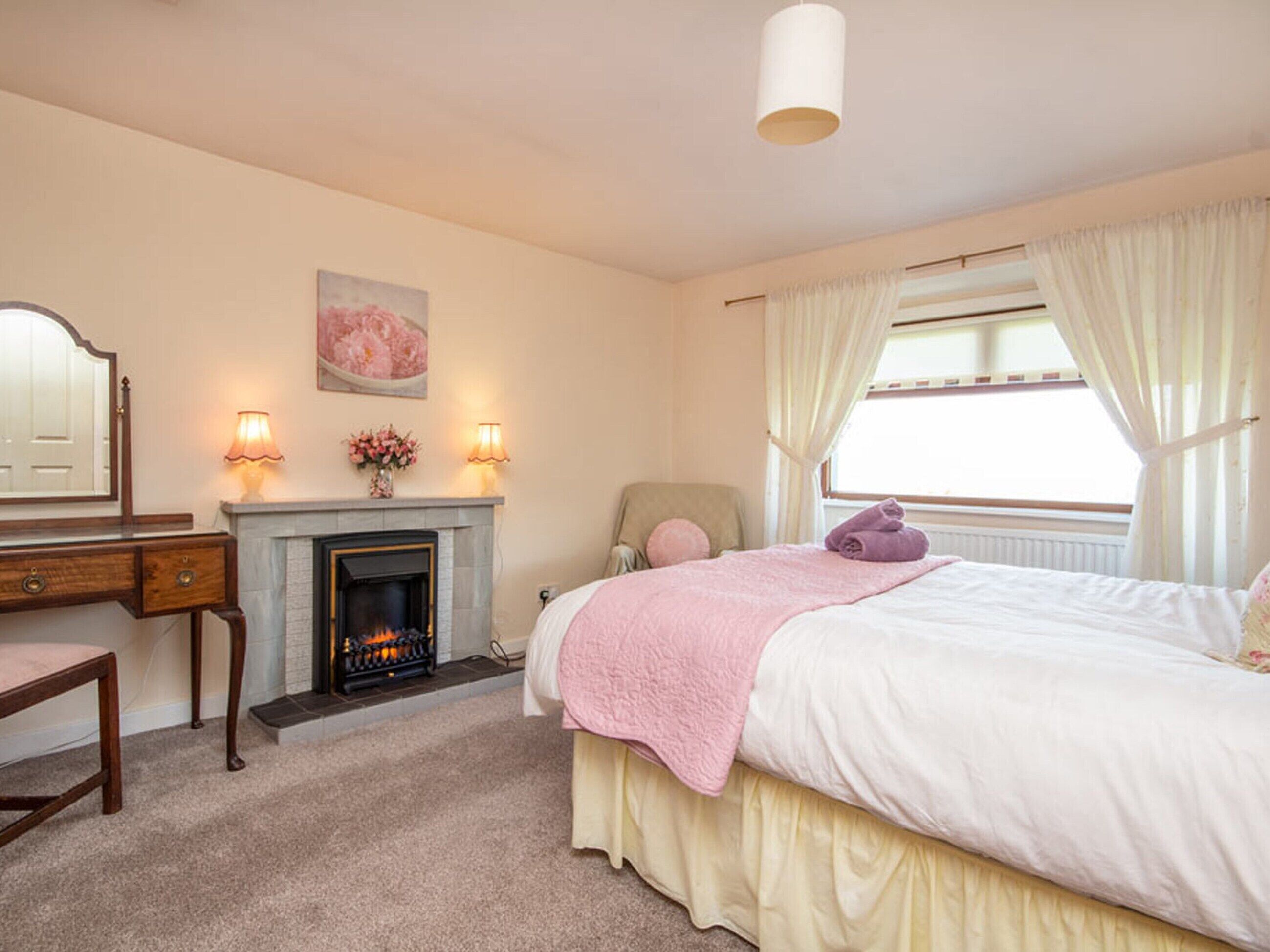 A bedroom with a fireplace, a bed with a pink blanket, a chair, and a desk with a mirror.