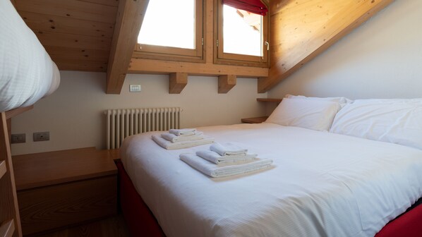 Une chambre avec un plafond en bois, un lit recouvert de draps blancs, un banc rouge et une fenêtre ornée d’un rideau rouge.