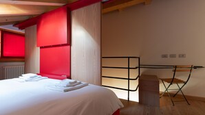 Quarto de hotel com uma cama, uma cadeira de madeira, um radiador e uma janela com cortinas vermelhas.