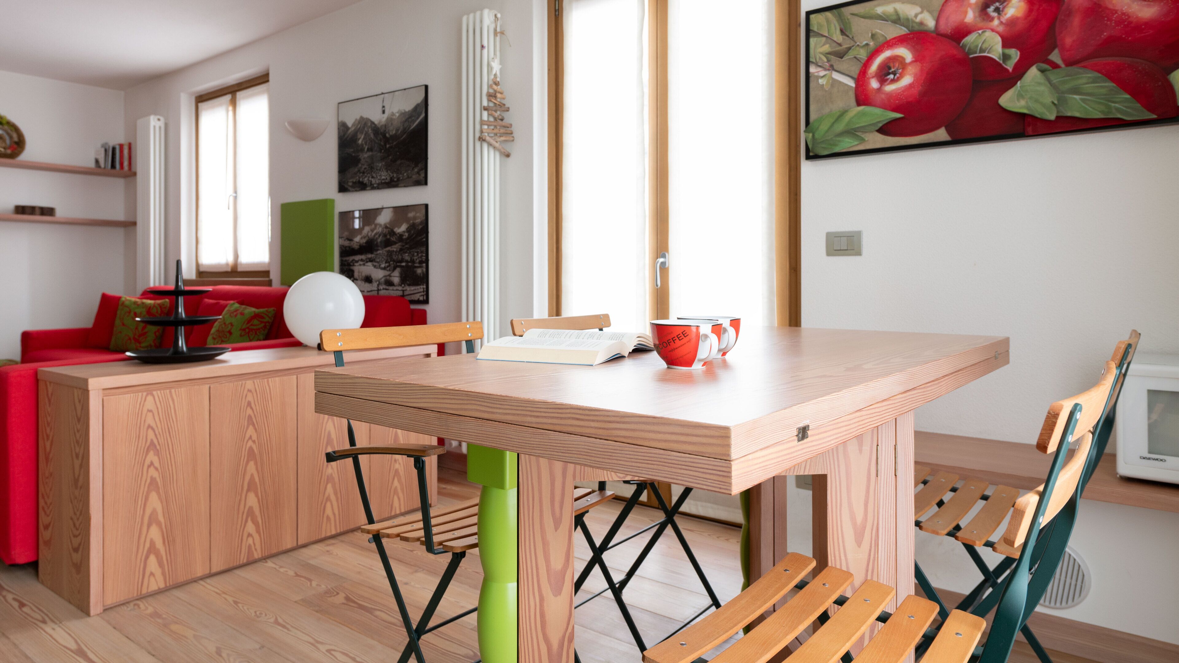 A modern kitchen with wooden cabinetry, a dining table, and chairs. There is a red couch in the background and a painting on the wall.