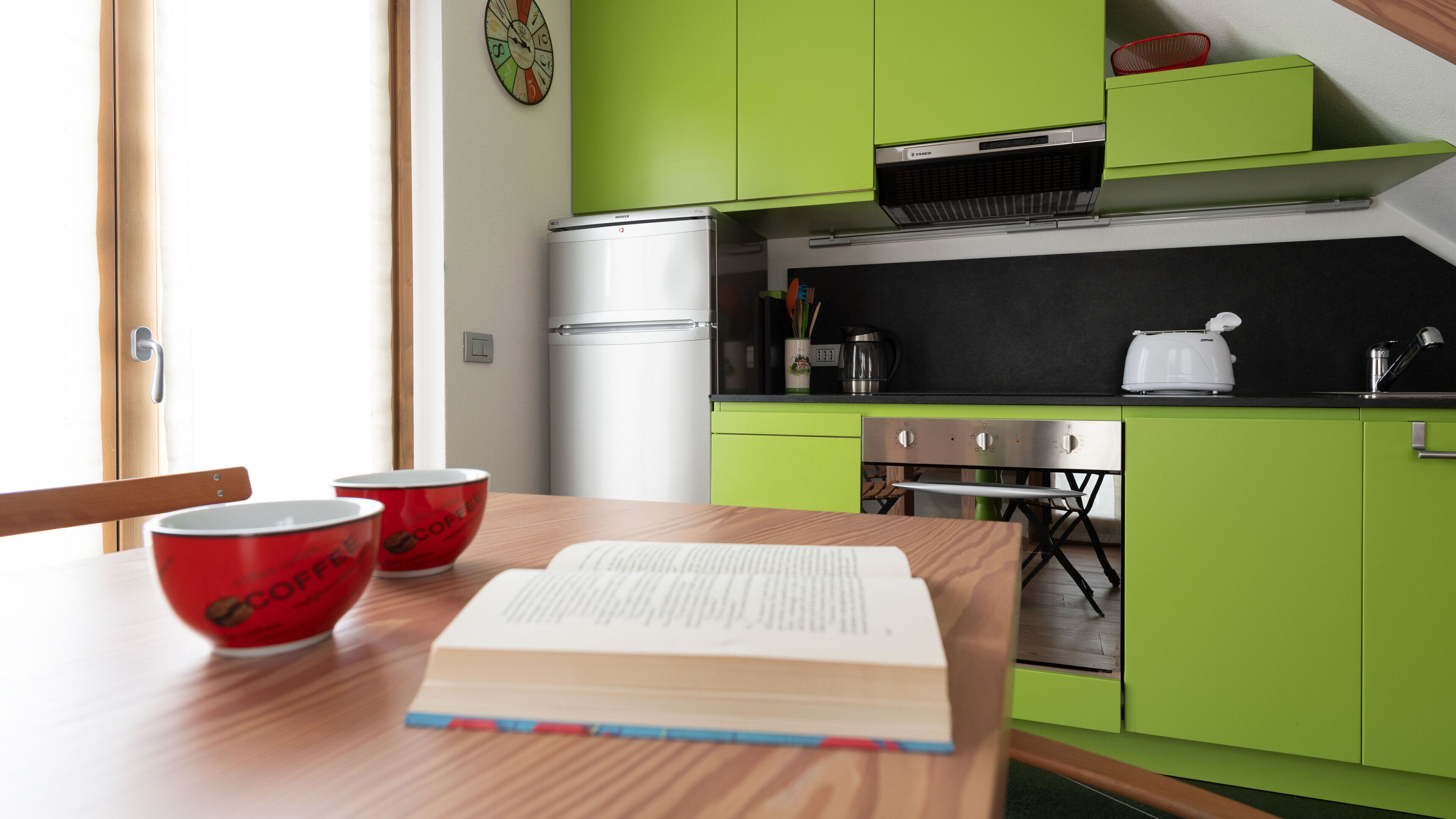 A compact kitchen with green cabinets, a stainless steel refrigerator, a black countertop, and a red coffee cup on a wooden table.