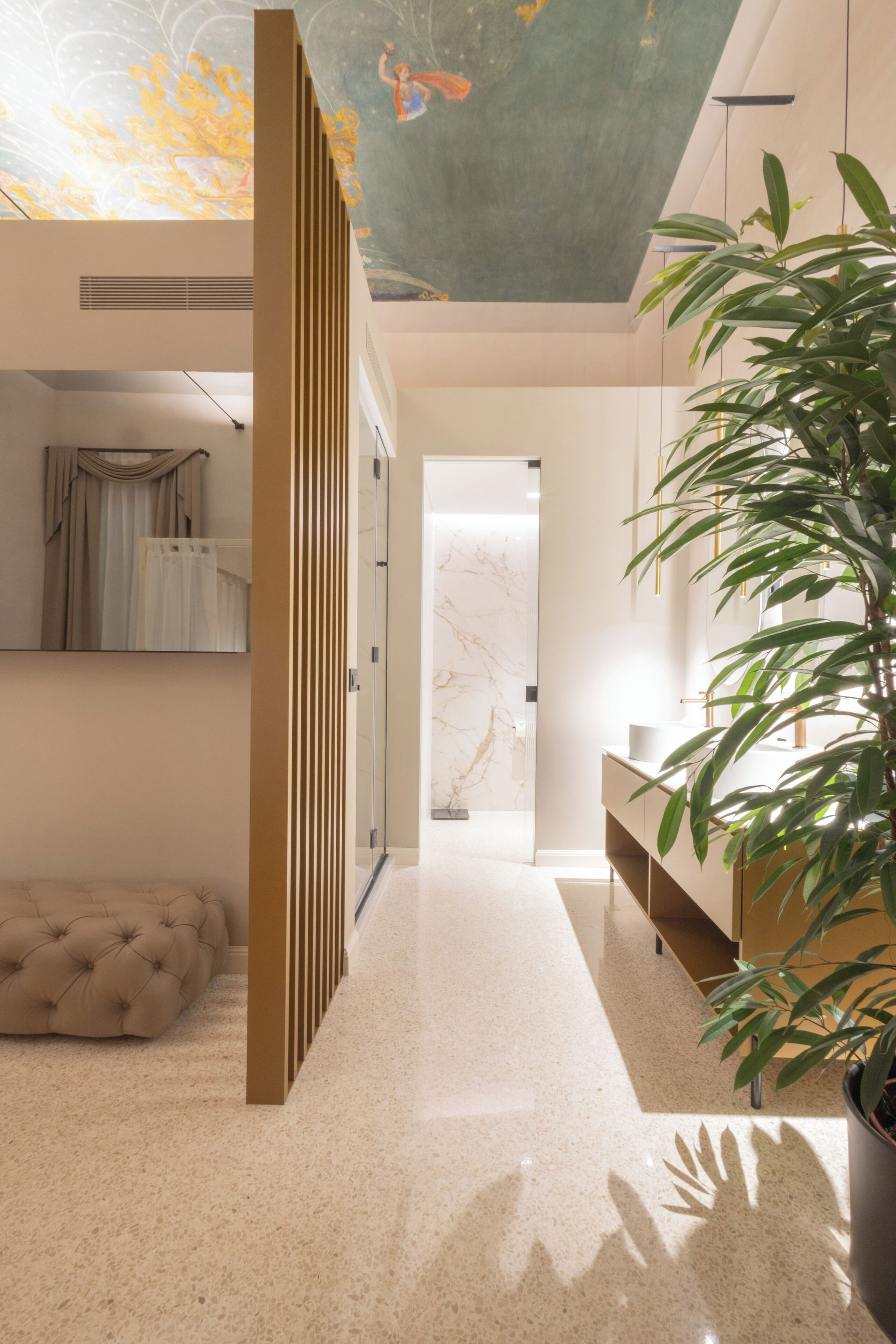 Un intérieur moderne avec un couloir menant à une salle de bain, comprenant un pouf capitonné, une plante en pot et un plafond orné d’une fresque.