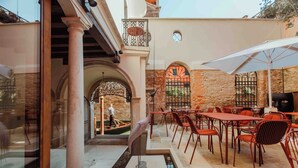 An outdoor patio with red metal chairs and tables, a white umbrella, and a stone wall with arched windows. - Palazzetto My Venice (Venice)