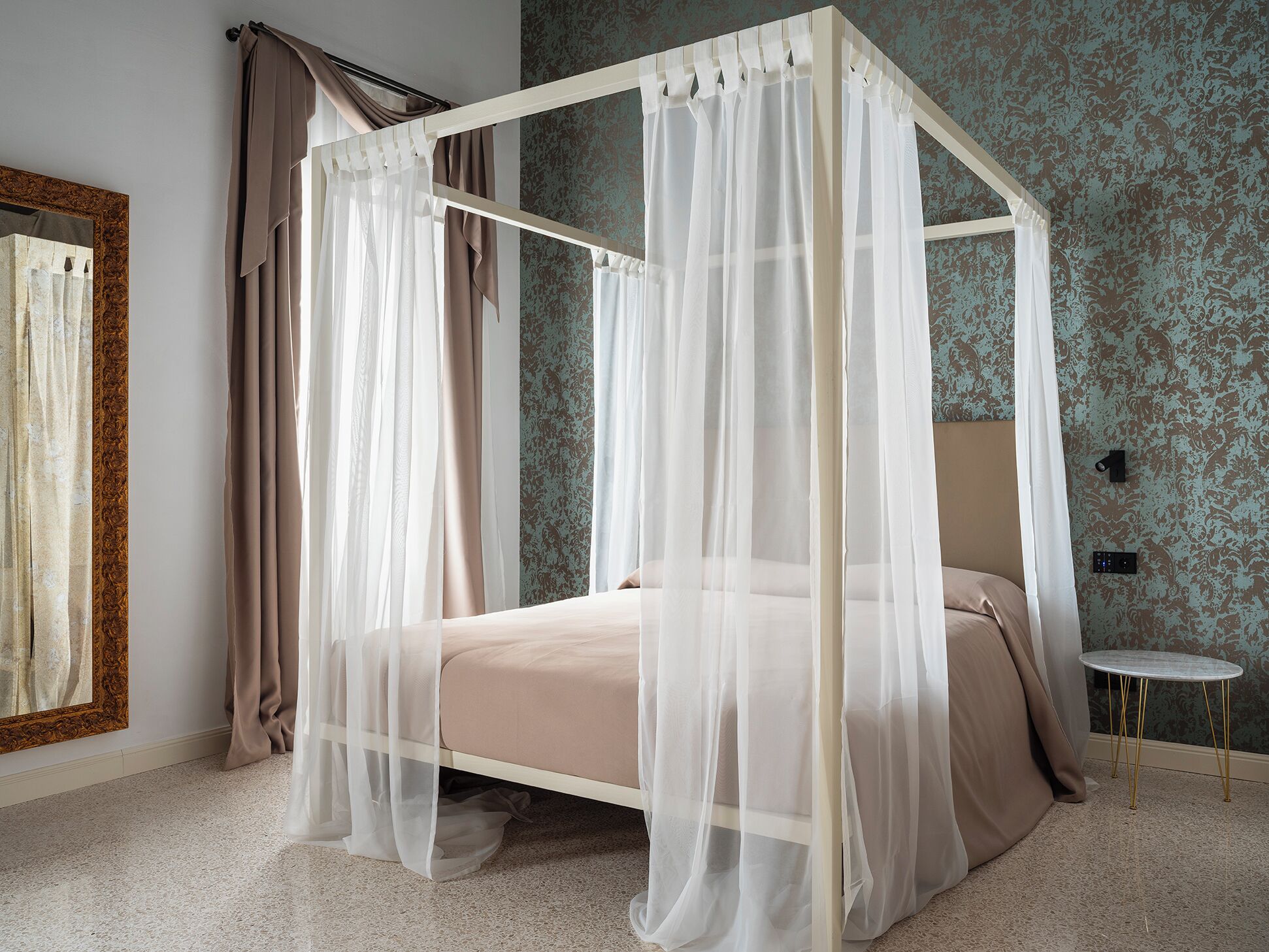 Una cama con dosel y cortinas translúcidas, colchón color beige y una mesita de noche con lámpara.
