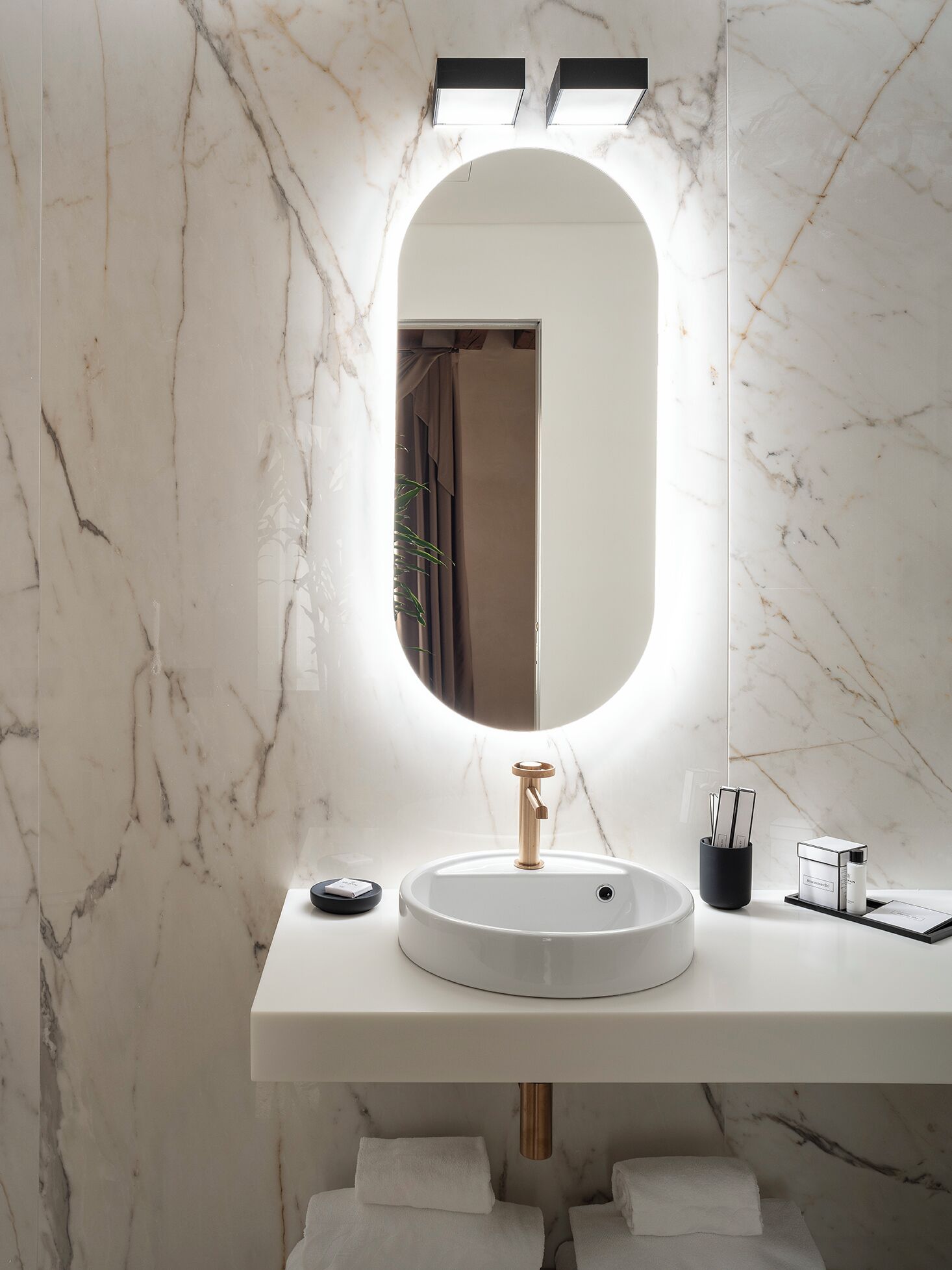A bathroom with a marble wall, a round sink, a mirror with integrated lighting, and a towel rack.