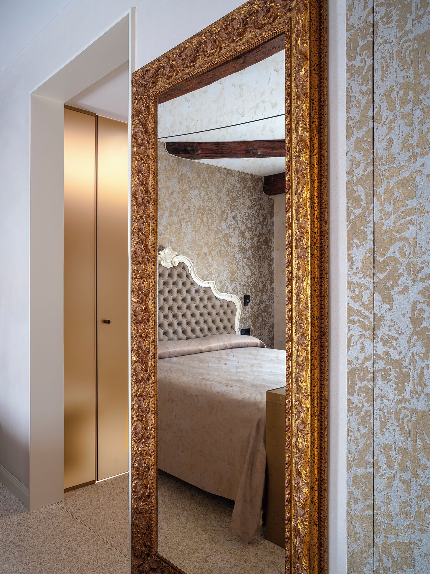 A bedroom with a large ornate mirror, a tufted headboard, and a patterned wallpaper.
