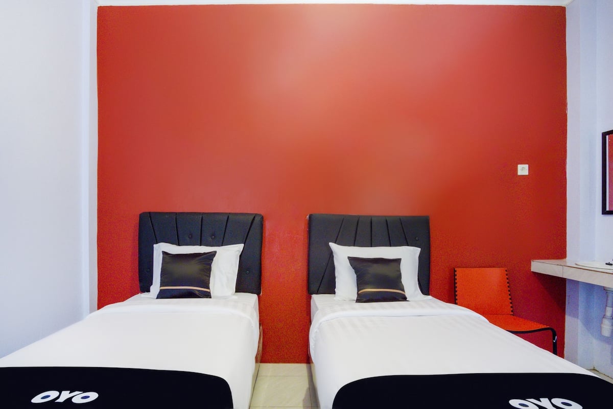 Une chambre d’hôtel avec deux lits, chacun doté d’une tête de lit noire et de draps blancs. Les murs sont peints en rouge, et on y trouve une petite chaise orange et une étagère blanche avec un miroir.