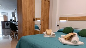 Ett modernt hotellrum med ett turkosfärgat sängöverkast, ett träskåp och utsikt över köksdelen.
