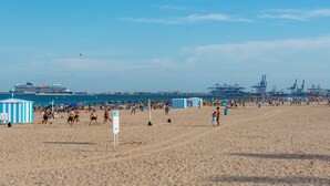 En strand där folk spelar volleyboll, ett fartyg i bakgrunden och industriella kranar.
