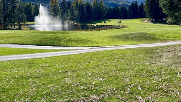 Un campo de golf con un estanque, árboles y montañas al fondo.