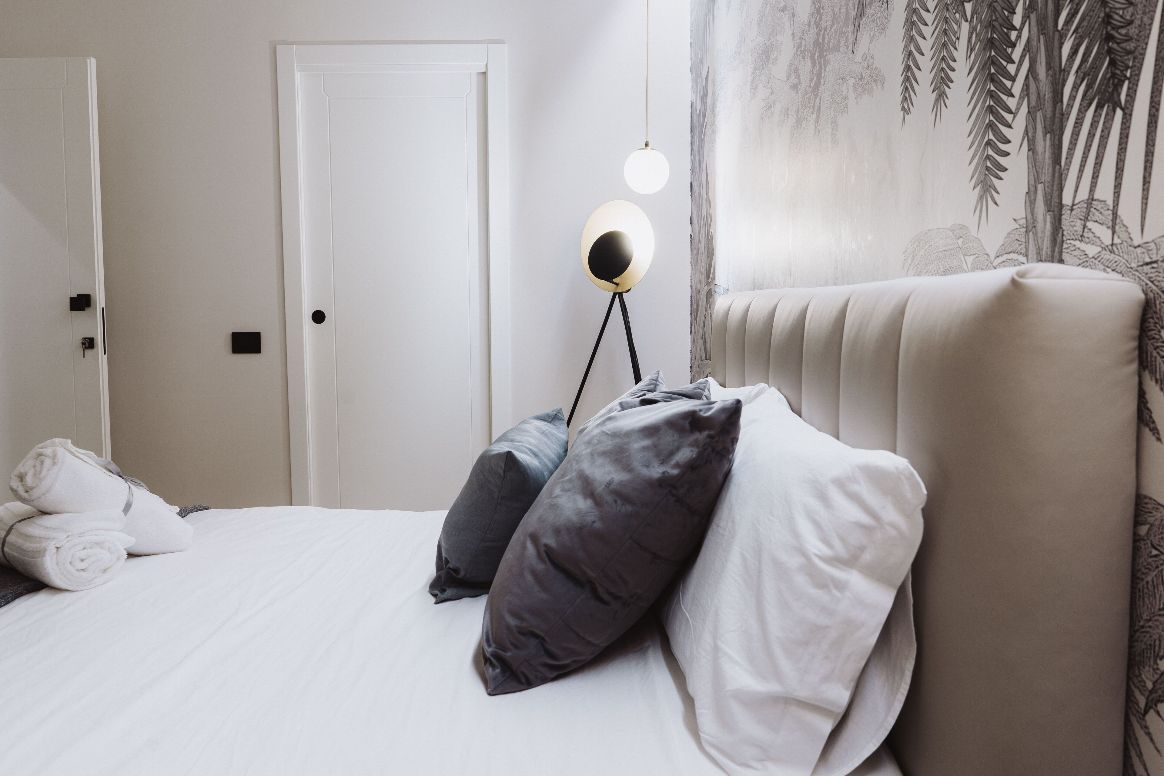 A neatly made bed with white and gray pillows, a round floor lamp, and a wall with a leaf pattern.
