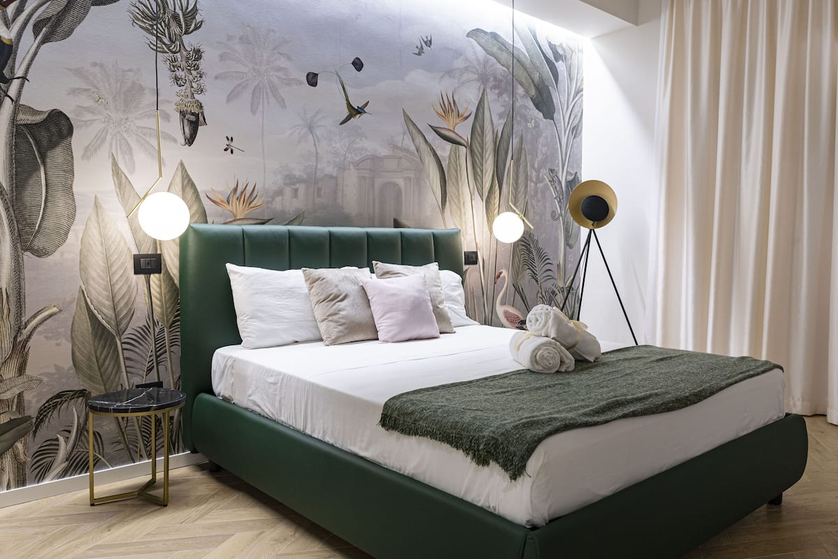 A bedroom with a green upholstered bed, white bedding, and a mural of tropical foliage on the wall. There are two bedside lamps and a small round table with a gold frame.