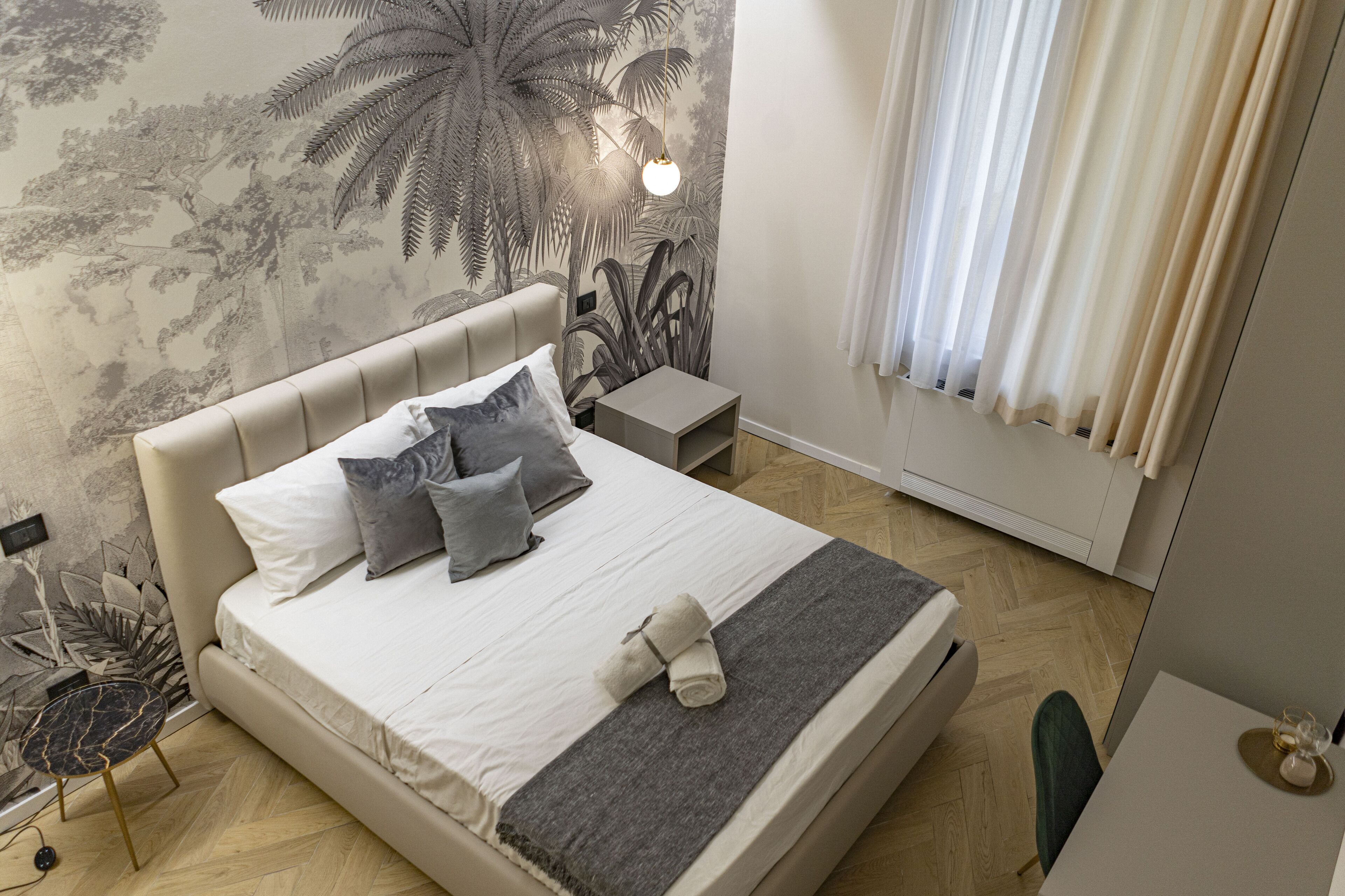 A neatly made bed with pillows, a bedside table, a lamp, and a wall with a tropical mural.