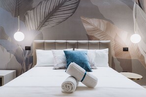 A neatly made bed with white and blue pillows, a rolled white towel, and a wall with a leaf pattern. - Ammare rooms (Agrigento)