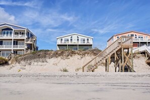 En ejendom ved stranden med et fleretages hus, altaner, en træterrasse og en sandstrand i forgrunden.