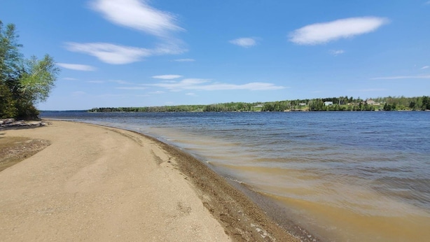 En sandstrand med en lugn flod, omgiven av träd och hus i fjärran.