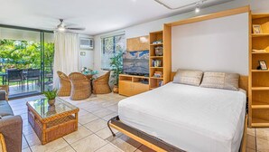 A modern bedroom with a large bed, a TV, a bookshelf, and a balcony with outdoor furniture. - 3-Min Walk to Beach! Ground Floor - No Resort Fee (Kihei)
