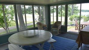 Un porche cubierto con una mesa redonda y bancos, ventanas amplias con vistas a un paisaje verde y un cuerpo de agua.
