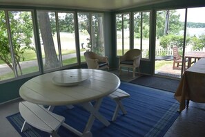 Un porche cubierto con una mesa redonda y bancos, ventanas amplias con vistas a un paisaje verde y un cuerpo de agua.