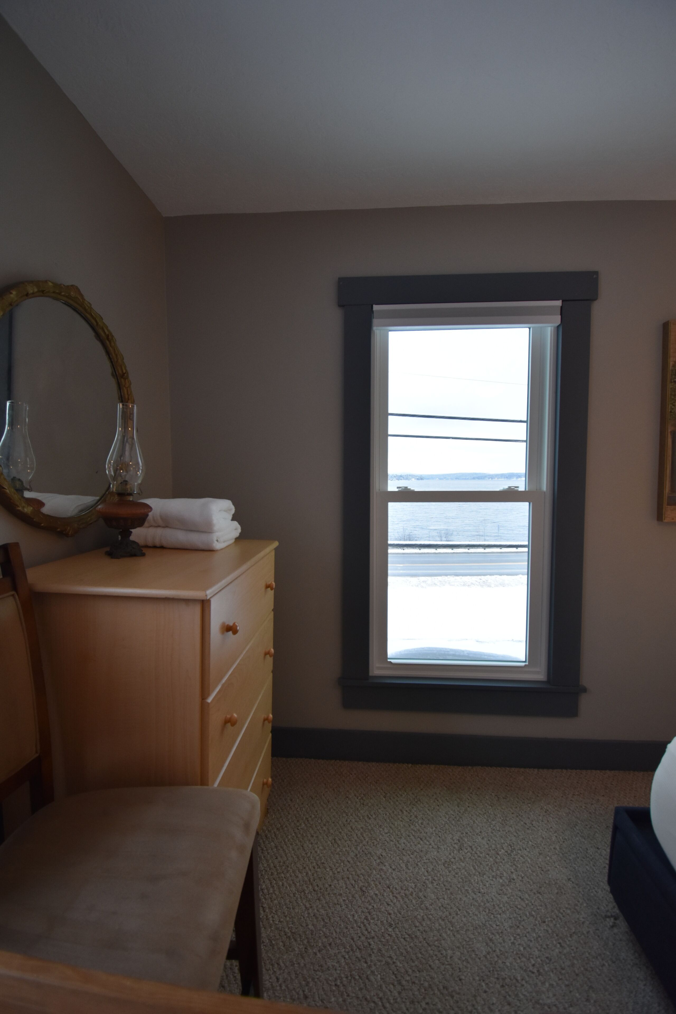 A bedroom with a large window, a wooden dresser, a mirror, and a bed.