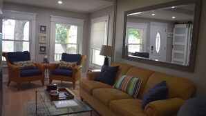 Ein Wohnzimmer mit einem gelben Sofa, zwei Korbstühlen mit blauen Kissen, einem Glastisch und einem großen Spiegel an der Wand.