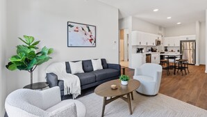 A modern living room with a grey sofa, a white armchair, and a coffee table. A large plant is in the corner, and a painting hangs on the wall. The kitchen is visible in the background with white cabinets and stainless steel appliances.