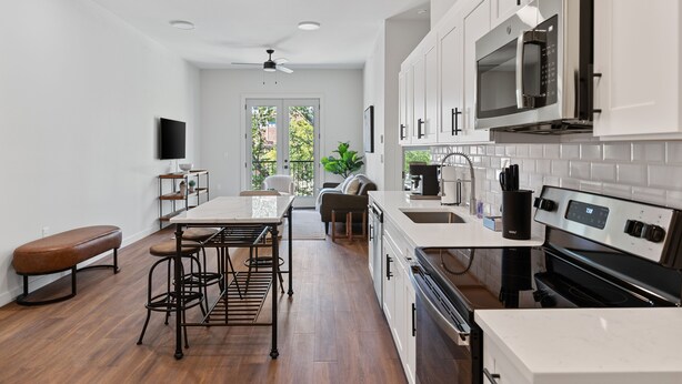 A modern kitchen with white cabinets, a stainless steel microwave, and a built-in oven. There is a dining area with a bench and stools, and a large window providing natural light.