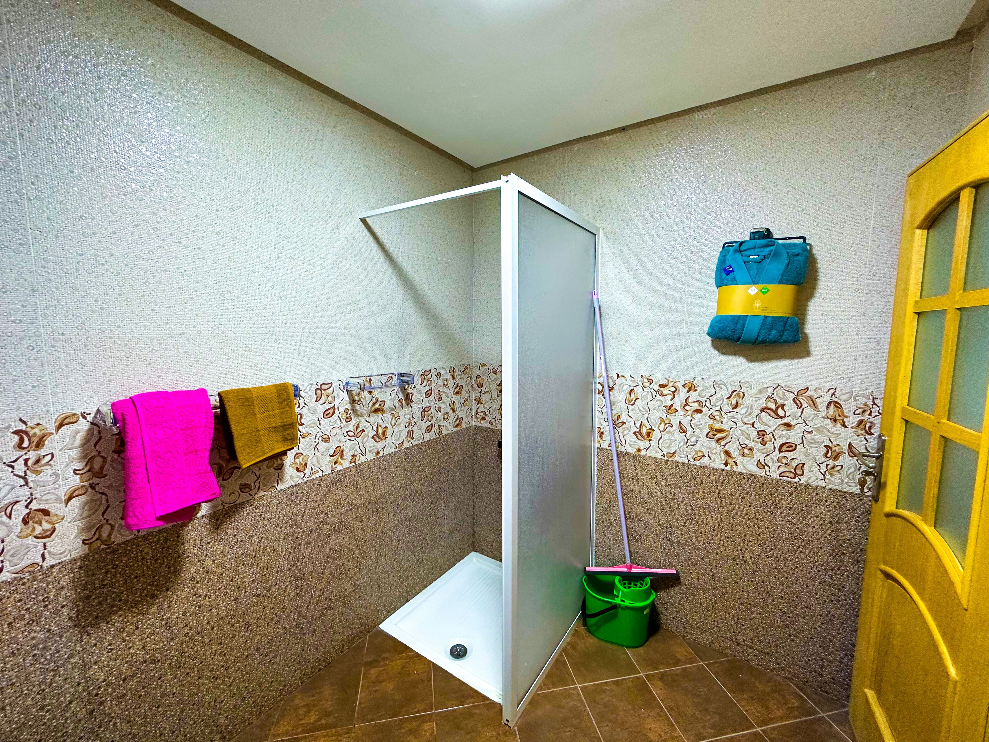 A small bathroom with a shower, a green bucket, a mop, and towels on the wall.
