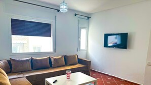A living room with a sofa, a coffee table, a TV, and a window with blinds. - Apartment with pool direct access to sea (Oued Laou)