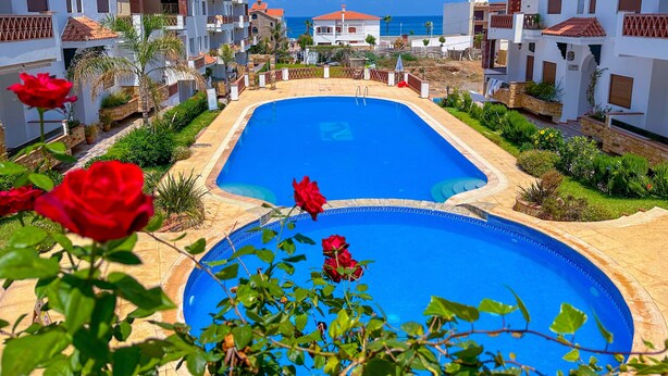 A swimming pool surrounded by white buildings with red-tiled roofs, a clear blue sky, and red roses in the foreground. - Apartment with pool direct access to sea (Oued Laou)