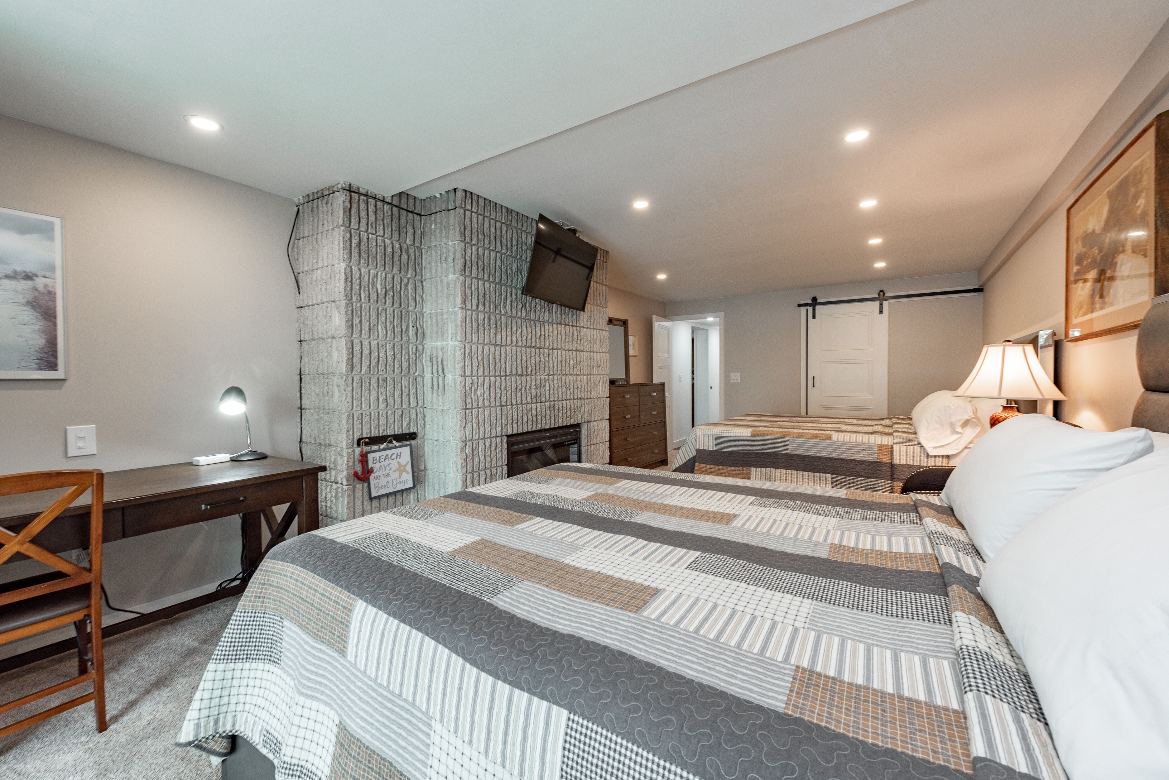 A bedroom with a large bed, a desk, a chair, a fireplace, and a door leading to another room.