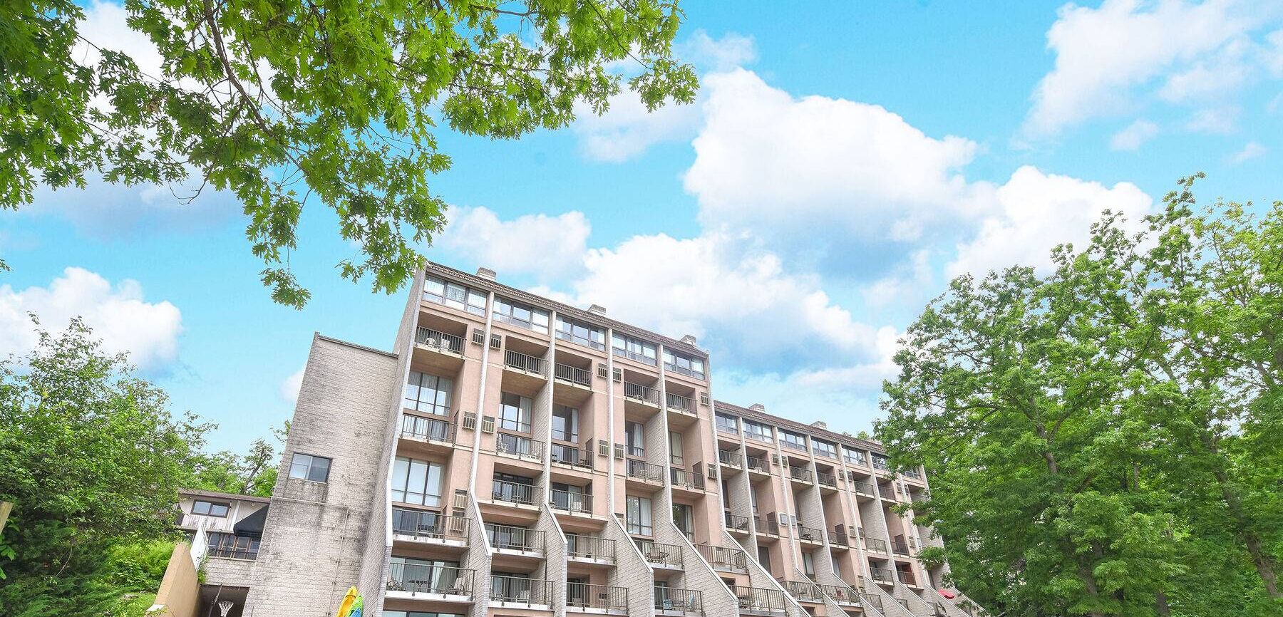 A multi-story residential building with balconies, surrounded by trees and a clear sky.