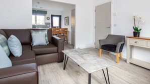Una sala de estar moderna con un sofá marrón, un sillón gris y una mesita. Se ve una cocina a través de un vano abierto.