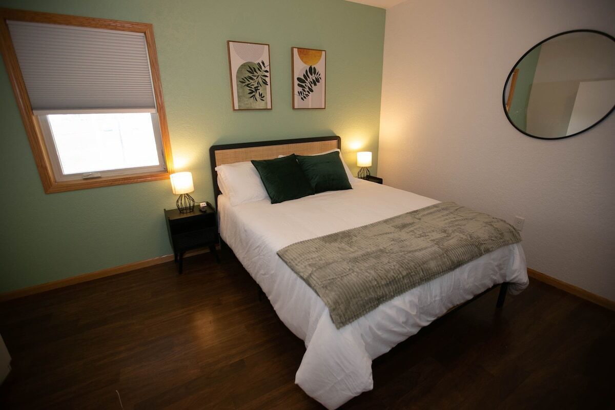 A neatly made bed with a white comforter and a green throw blanket, flanked by two bedside lamps, in a room with wooden flooring and a window with blinds.