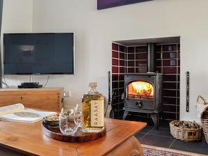 Una sala de estar con televisor, una mesa de madera con una botella, vasos y un libro, además de un estufa de leña.