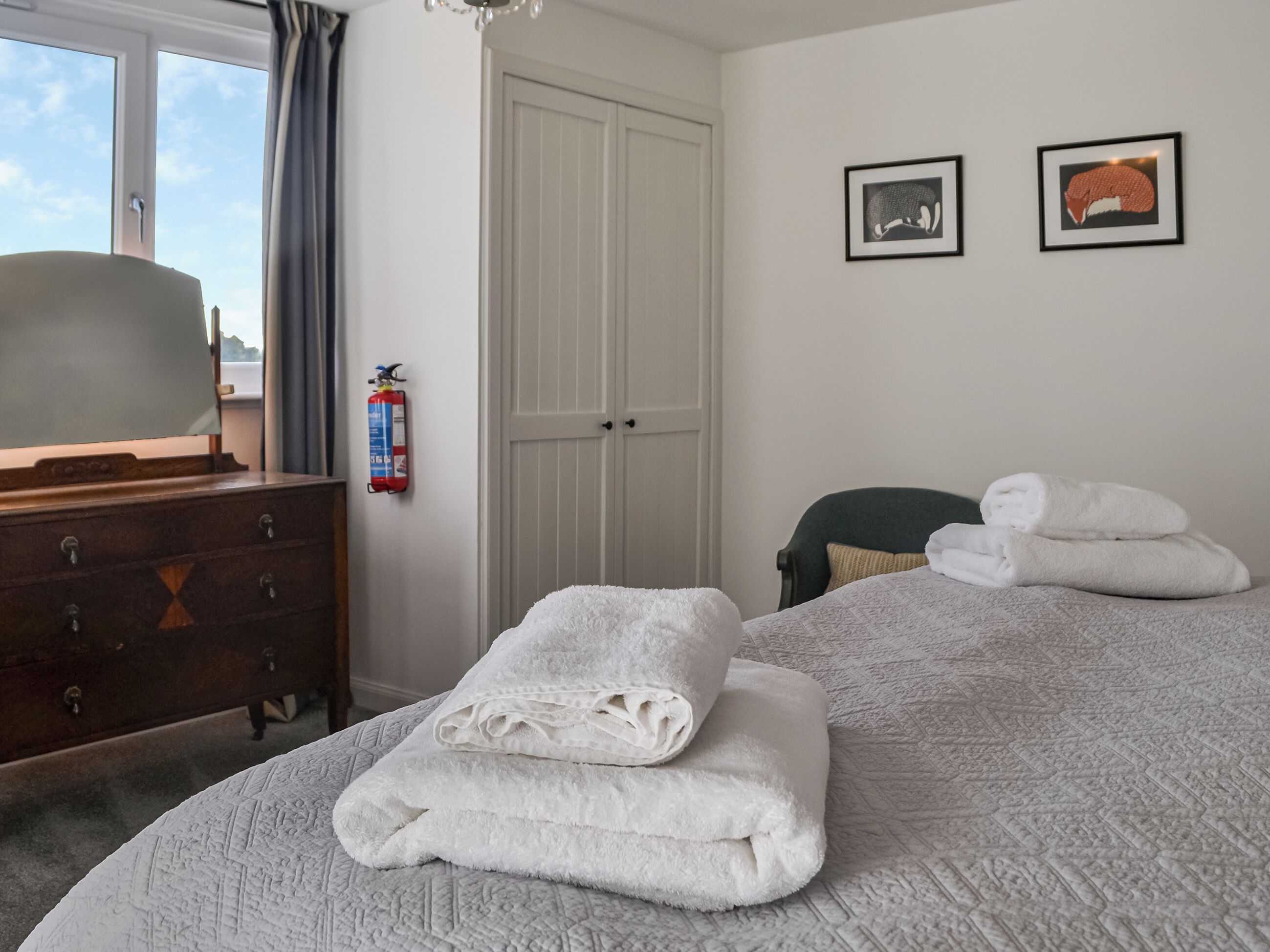 Ein Schlafzimmer mit einem Bett, einem Schminktisch, einem Feuerlöscher und zwei gerahmten Bildern an der Wand.