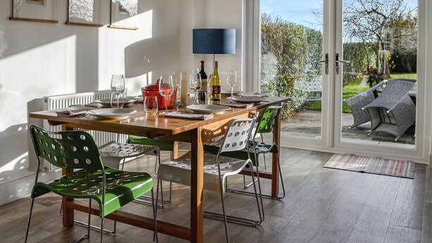 A dining area with a wooden table, green chairs, and a view of the garden through glass doors.