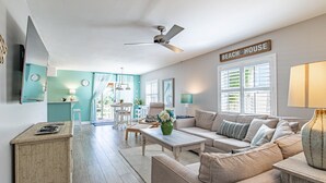 En stue med sofa, sofabord og loftventilator. Der er et skilt på væggen, der står "BEACH HOUSE".
