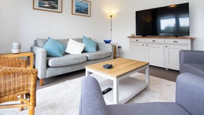A living room with a grey sofa, blue cushions, a wooden coffee table, a wicker chair, and a flat-screen TV on a white cabinet.