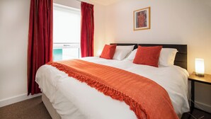 A neatly made bed with a white and orange bedspread, red curtains, a framed picture on the wall, and a bedside table with a lamp.
