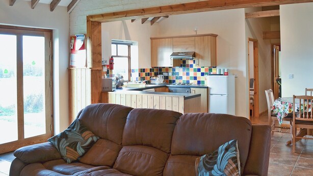 A living room with a brown leather sofa, wooden beams on the ceiling, and a kitchen in the background.