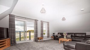A spacious living room with a large black leather sofa, a wooden coffee table, and a TV on a wooden stand. There are two windows with grey curtains, and a view of a beach is visible through the windows. - Amazing coastal location with access to sandy beach. (Balintore, near Tain)