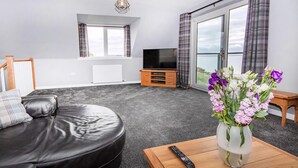 A modern living room with a leather sofa, a wooden coffee table with a vase of flowers, a TV on a wooden stand, and large windows with curtains. - Amazing coastal location with access to sandy beach. (Balintore, near Tain)