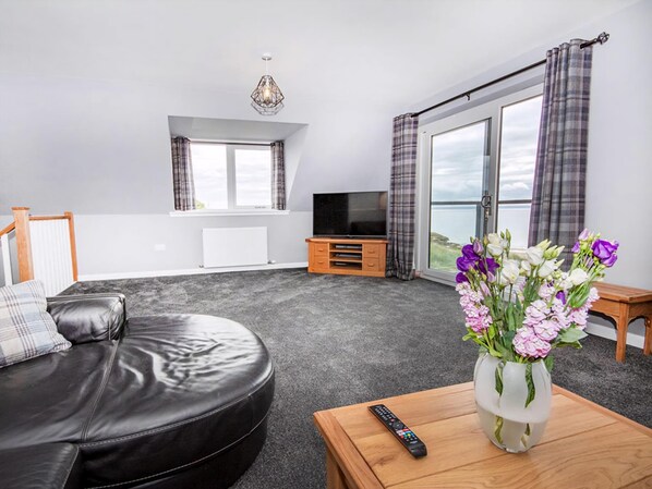 A modern living room with a leather sofa, a wooden coffee table with a vase of flowers, a TV on a wooden stand, and large windows with curtains. - Amazing coastal location with access to sandy beach. (Balintore, near Tain)