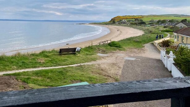 A coastal view with a sandy beach, a wooden bench, and a path leading to a house with a white fence. - Amazing coastal location with access to sandy beach. (Balintore, near Tain)