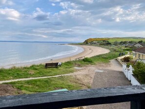 A coastal view with a sandy beach, a wooden bench, and a path leading to a house with a white fence. - Amazing coastal location with access to sandy beach. (Balintore, near Tain)