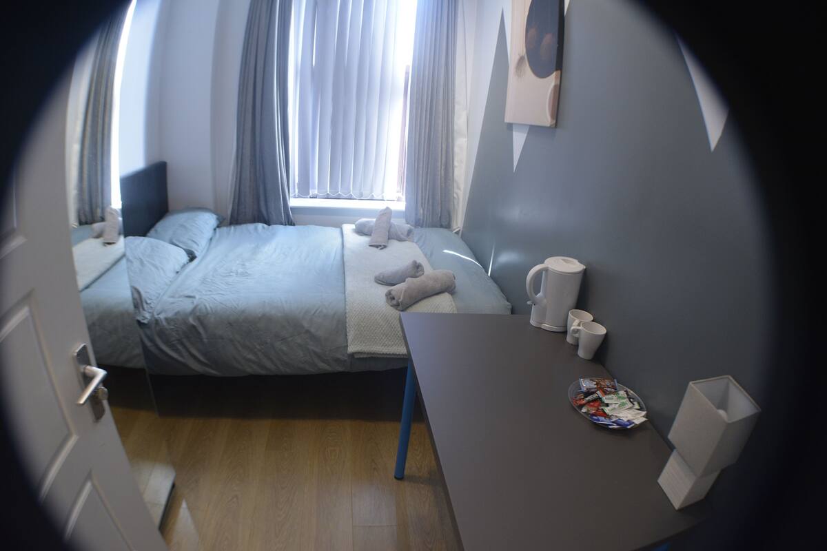 A compact hotel room with a bed, a small table, a chair, a window with curtains, and a wall-mounted shelf with items.
