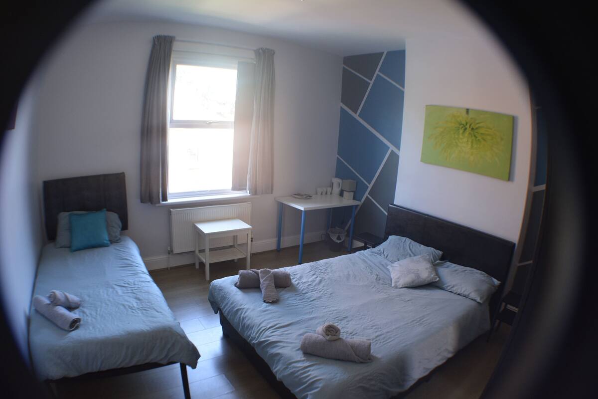 A hotel room with two single beds, a desk, a chair, a radiator, and a window with curtains.